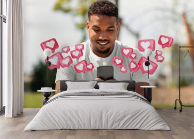 A young black man is sitting in a park, smiling while looking at his smartphone. He is holding the phone with both hands, and there are multiple heart-shaped icons floating around him Wall mural