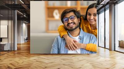 A young Indian couple shares a cheerful embrace on a comfortable sofa, showcasing their affection. The inviting indoor environment and joyful expressions highlight their connection. Wall mural