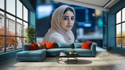 A young woman wearing a headscarf is focused on coding at her desk, surrounded by multiple screens displaying programming code in a contemporary office setting Wall mural