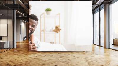 African American father and daughter are enjoying a special bonding moment in their cozy living room. The girl, smiling widely, is playfully hugging her dad from behind while he sits comfortably Wall mural