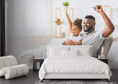 African American father and his daughter are sitting on a couch together watching television. They are both smiling and seem to be enjoying their movie night, copy space Wall mural