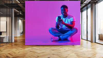 African American guy sitting cross legged with smartphone, surfing social media, chatting to friend in neon light Wall mural