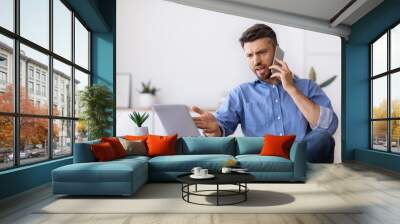 Angry businessman talking on cellphone and looking at laptop screen in office Wall mural