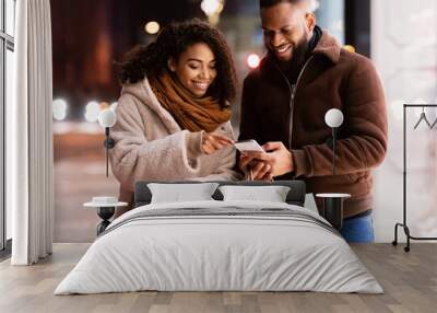 App For Online Shopping. Portrait of smiling black man and woman using mobile phone, lady pointing at screen, couple holding shopper bags, standing outdoors in the evening, choosing gift Wall mural