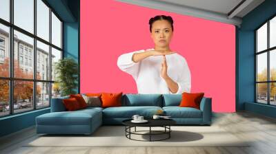 Asian woman is performing a stop gesture with her hands, expressing authority and confidence against a bright pink background. Wall mural