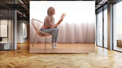 Attractive African American woman in hijab reading fascinating book in cozy armchair at home, blank space Wall mural