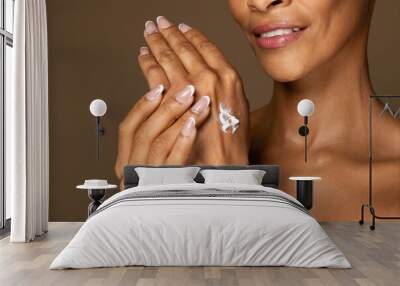 Attractive black middle aged woman applying moisturising lotion on hands, making beauty routine, brown background Wall mural