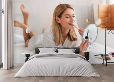 Attractive middle aged woman applying lipstick, looking at mirror while lying on bed and putting on decorative makeup Wall mural