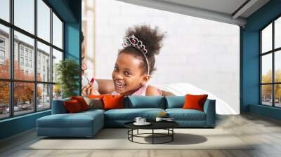 Black father and his daughter share a delightful moment, surrounded by light and warmth in their living room. The little girl wears a playful tiara and smiles brightly as she engages with her dad Wall mural