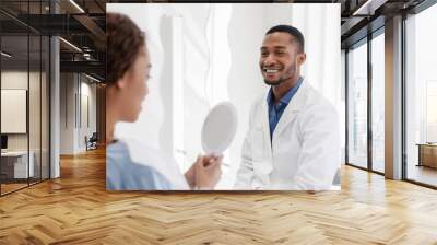 Black lady checking her white smile in mirror after stomatological treatment in modern clinic, happy dentist watching patient Wall mural