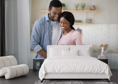 Black pregnant woman and her handsome husband embracing and smiling while spending time together near window at home Wall mural