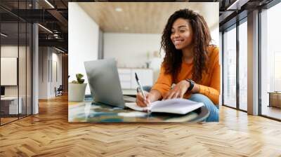 Black woman sitting at desk, using computer writing in notebook Wall mural