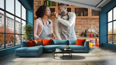 Black young man feeding his beautiful woman while cooking together at kitchen, empty space Wall mural