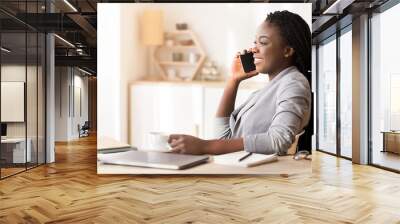 Break at work. Smiling african american businesswoman talking on cellphone and drinking coffee, relaxing at workplace Wall mural