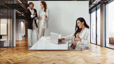 Businesswomen pointing at pregnant woman working on laptop in office, worker harassment and discrimination Wall mural