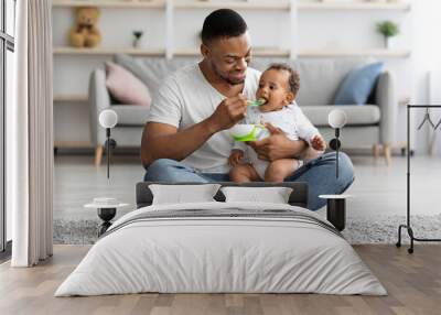 Caring Black Dad Feeding His Adorable Infant Baby From Spoon At Home Wall mural