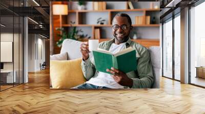 Cheerful adult african american man in glasses and casual reading book and enjoy favorite drink on sofa Wall mural