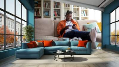 Cheerful african man using phone and credit card at home Wall mural