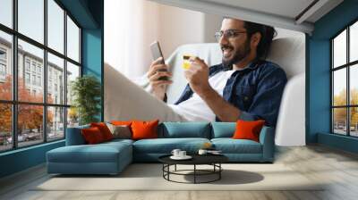 Cheerful Arab Guy Shopping Online With Smartphone And Credit Card At Home Wall mural