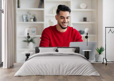 Cheerful attractive millennial arabic businessman with beard in red clothes typing on laptop in home office Wall mural