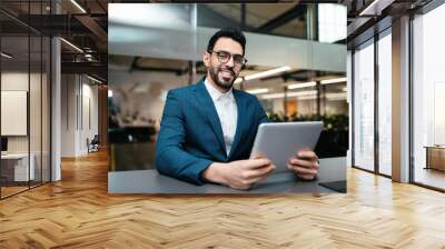 Cheerful attractive young middle eastern male with beard in glasses and suit sits at table with tablet in office Wall mural