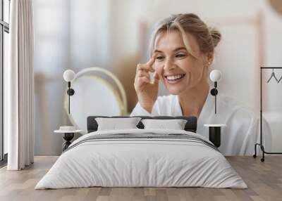 Cheerful beautiful blonde middle aged lady in silky bathrobe using moisturizing eye cream, looking at mirror and smiling, enjoying face care routine, closeup shot. Dark circles, eye wrinkles treatment Wall mural