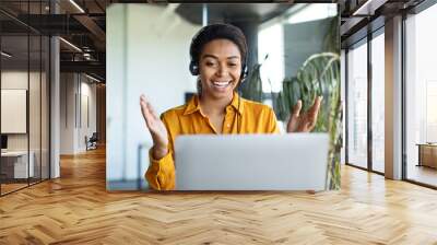 Cheerful black female manager making video call wearing headset and talking to laptop webcam sitting in office Wall mural