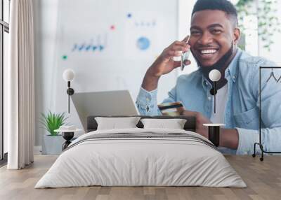 Cheerful black guy talking on phone and holding credit card Wall mural