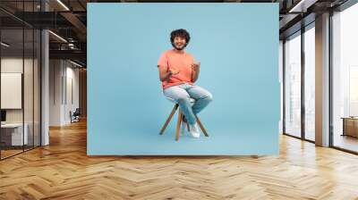 Cheerful eastern guy sitting on chair over blue background Wall mural