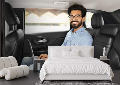 Cheerful eastern guy using laptop while sitting in taxi Wall mural