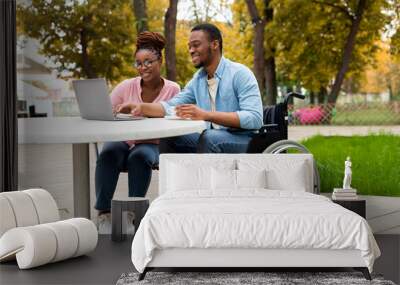 Cheerful impaired black man with female colleague using laptop together in outdoor cafe at autumn park Wall mural