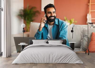 Cheerful indian man with laptop communicating on phone at home Wall mural