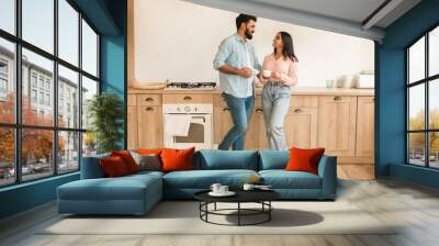 Cheerful loving indian couple enjoying morning coffee at home, happy spouses having conversation, standing in kitchen Wall mural