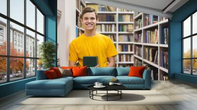 Cheerful male student standing between bookshelves in library Wall mural