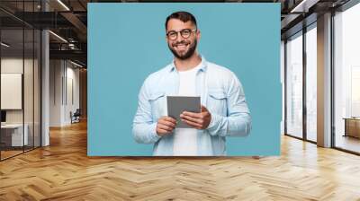 Cheerful mature caucasian guy in glasses with tablet look at camera, isolated on blue background, free space Wall mural