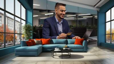 Cheerful middle eastern entrepreneur using laptop, holding credit card Wall mural