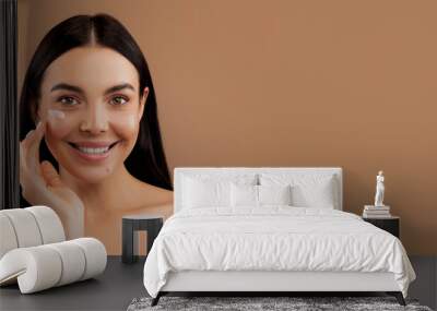 Cheerful sensual naked long-haired brunette millennial woman with smile on her face using facial eye cream for young smooth glowing skin, isolated on beige studio background, copy space Wall mural