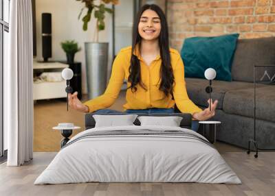 Cheerful spanish lady sitting on floor by couch, meditating with closed eyes and smiling at home, living room interior Wall mural