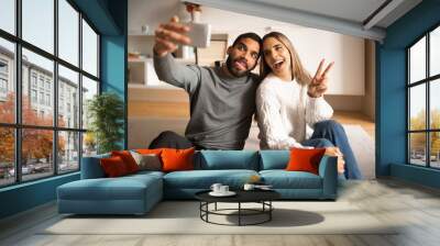 Cheerful young arab man hugging european wife, make peace gesture and photo on smartphone Wall mural