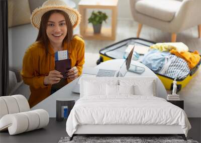 Cheerful young lady with passport and ticket using laptop to buy tourist vacation at online travel agency from home Wall mural