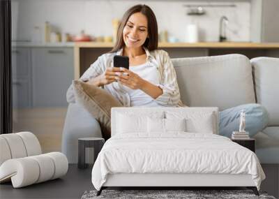 Cheerful young woman texting on her smartphone, sitting cross-legged on comfortable couch Wall mural