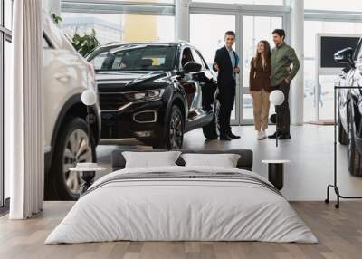 Confident salesman showing new car to young Caucasian couple at auto dealership Wall mural