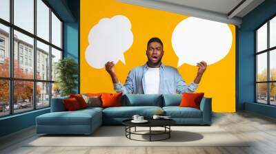 Confused black guy holding two speech bubbles on yellow background Wall mural