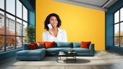 Consumerism. Charming afro girl holding credit card, thinking about purchase, what to buy. Copyspace, yellow wall Wall mural