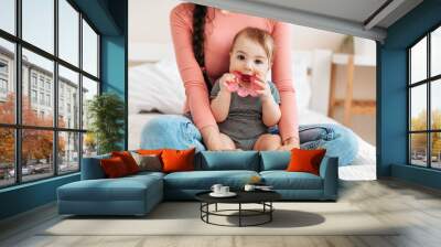 Cute little baby girl playing with teething toy, biting teether and looking at camera, sitting with mother on bed at home Wall mural