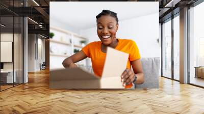 Delivery, remote shopping. Smiling black woman unboxing cardboard package, happy with her purchase at home Wall mural