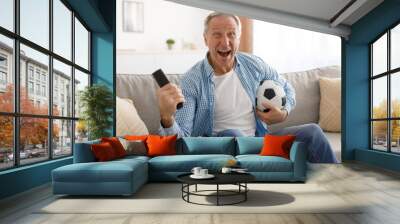 Excited mature man watching tv celebrating goal holding ball Wall mural