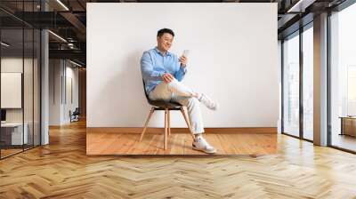 Excited middle aged asian man using smartphone, networking in social media, sitting on chair over white studio wall Wall mural