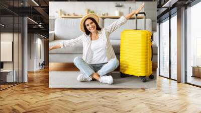 Excited woman in straw hat, sitting with arms wide open beside suitcase Wall mural