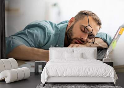 Exhausted millennial man sleeping on his office desk, next to laptop and documents, tired of overworking Wall mural